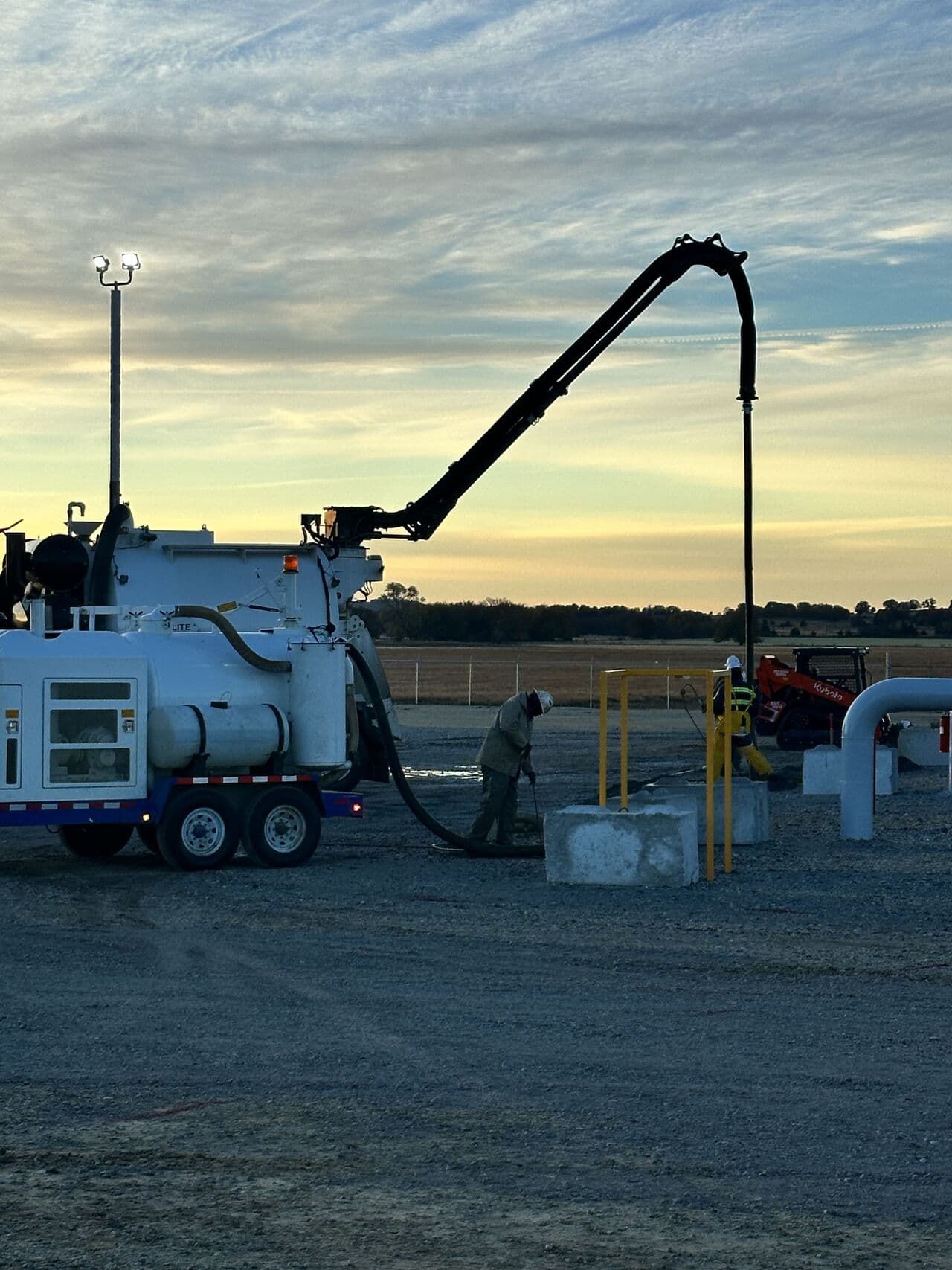 A vacuum excavation truck extending its boom over a pipeline site at dusk while a crew member works below