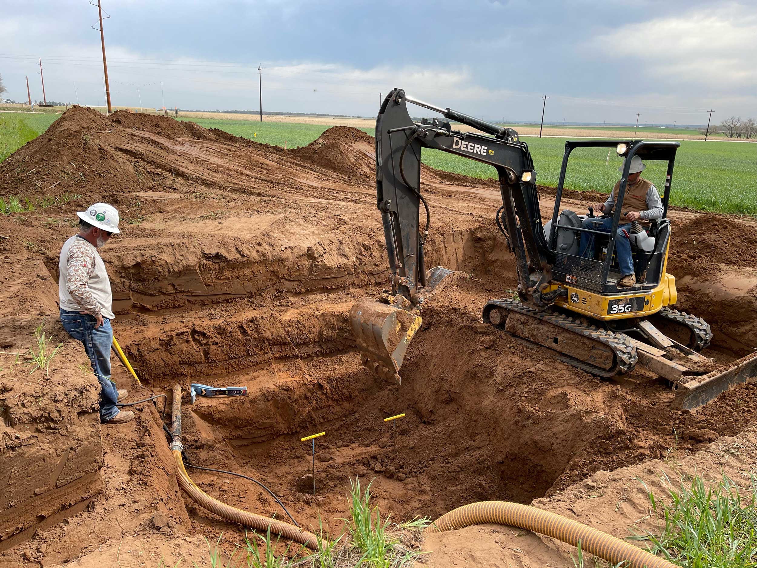Two crew members and a compact excavator working in an open trench cut through farmland