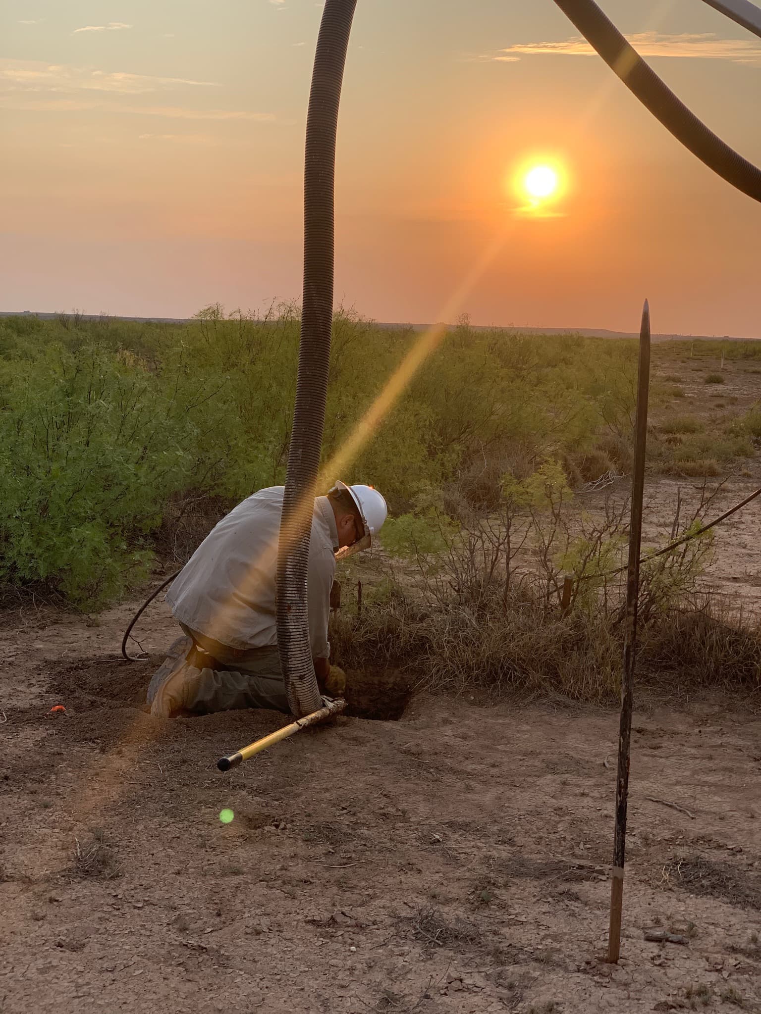 A hard-hatted worker kneeling to guide a vacuum hose into a small excavation at sunset